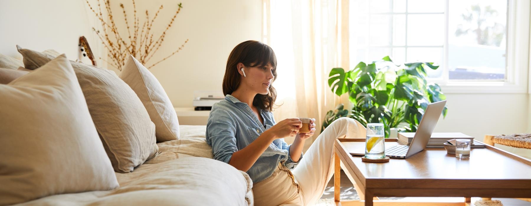woman holds a cup of coffee and watches her laptop as she sits in her sun filled living room
